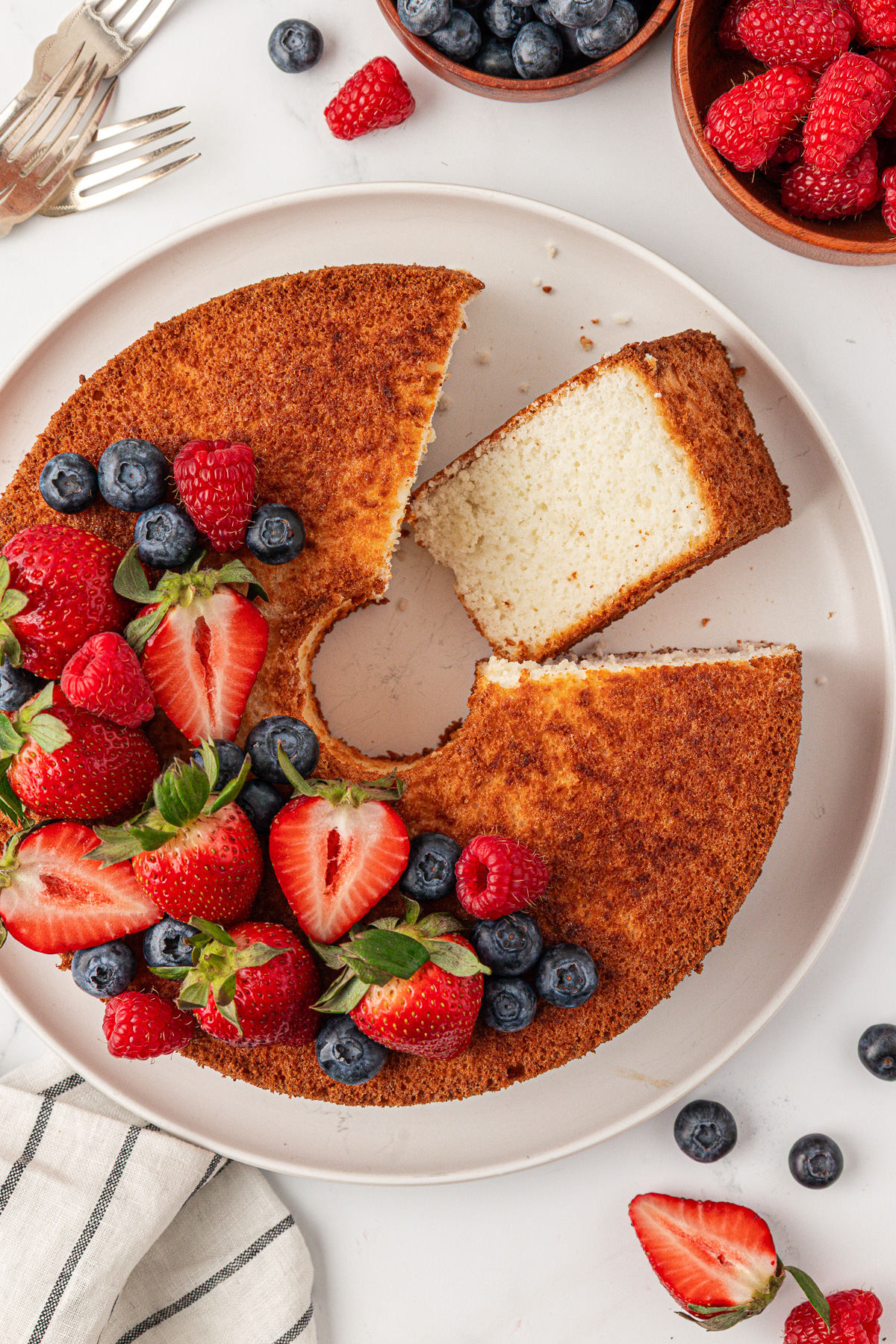 A round, golden-brown angel food cake on a white plate, topped with fresh strawberries, blueberries, and raspberries. One slice of the fluffy angel food cake is cut and slightly pulled out. Bowls of berries and a striped cloth are nearby.