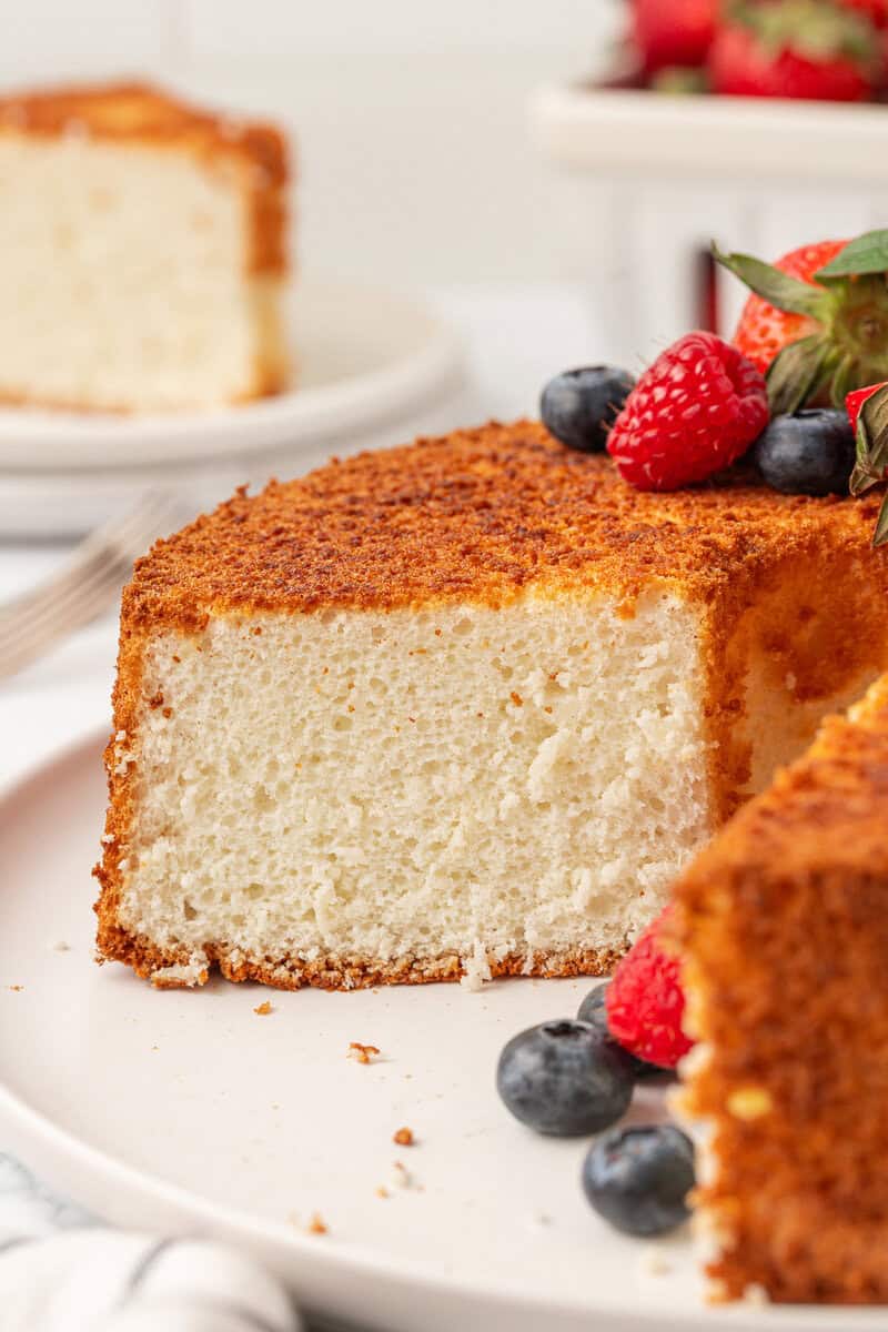 A close-up of a fluffy slice of angel food cake on a white plate, beautifully garnished with fresh strawberries, raspberries, and blueberries. Another light and airy slice of angel food cake appears in the background.