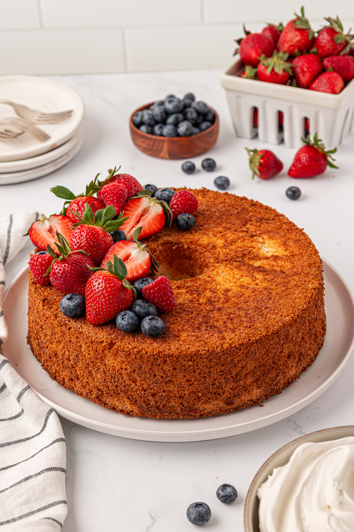 A golden angel food cake on a white plate, topped with fresh strawberries and blueberries. In the background, more strawberries fill a basket, alongside plates, a bowl of blueberries, and a dish of whipped cream.