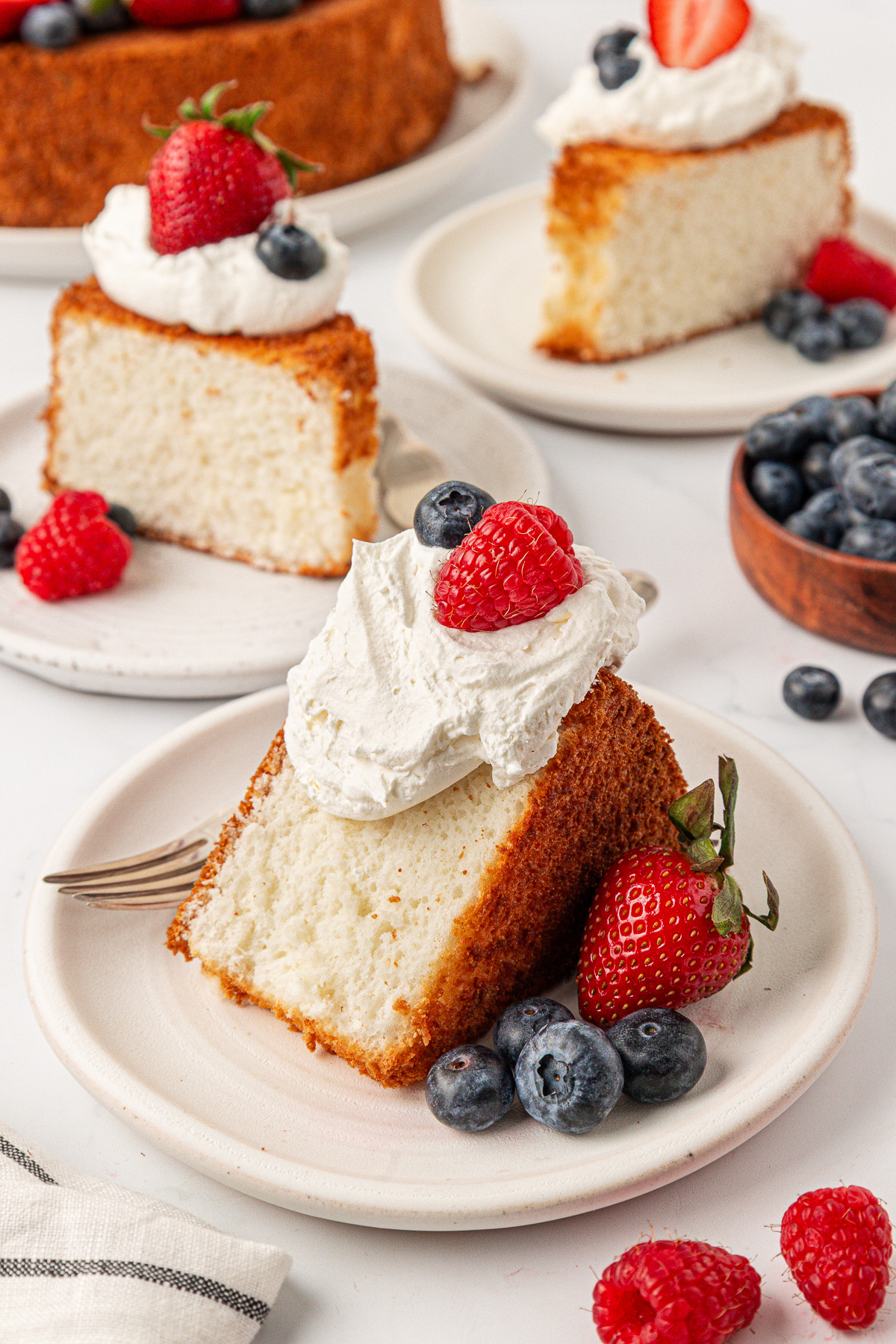 A slice of angel food cake topped with whipped cream, a raspberry, and blueberries sits on a plate, surrounded by fresh berries. Additional slices of angel food cake and more berries are visible in the background.
