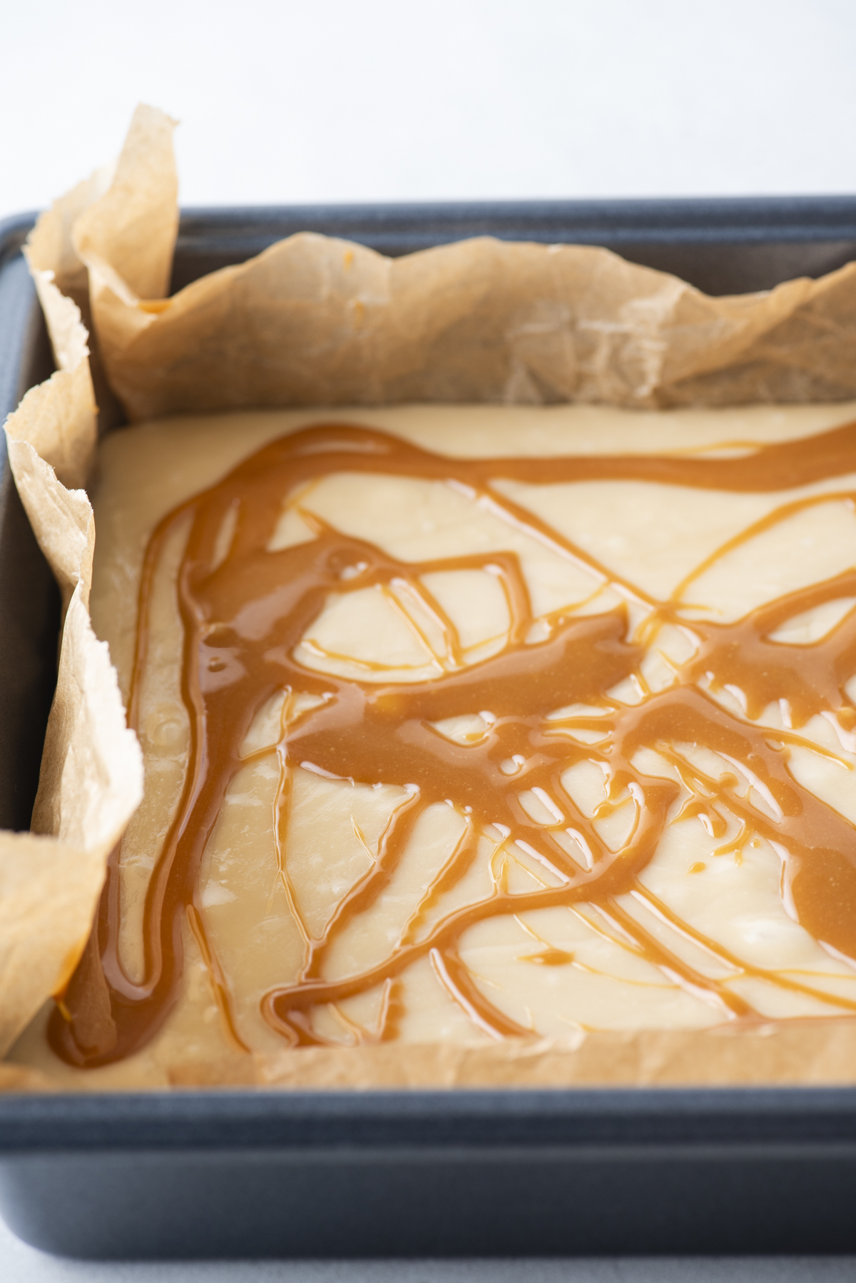 A baking pan lined with parchment paper holds a creamy caramel apple fudge dessert topped with swirls of caramel sauce, ready to be baked.