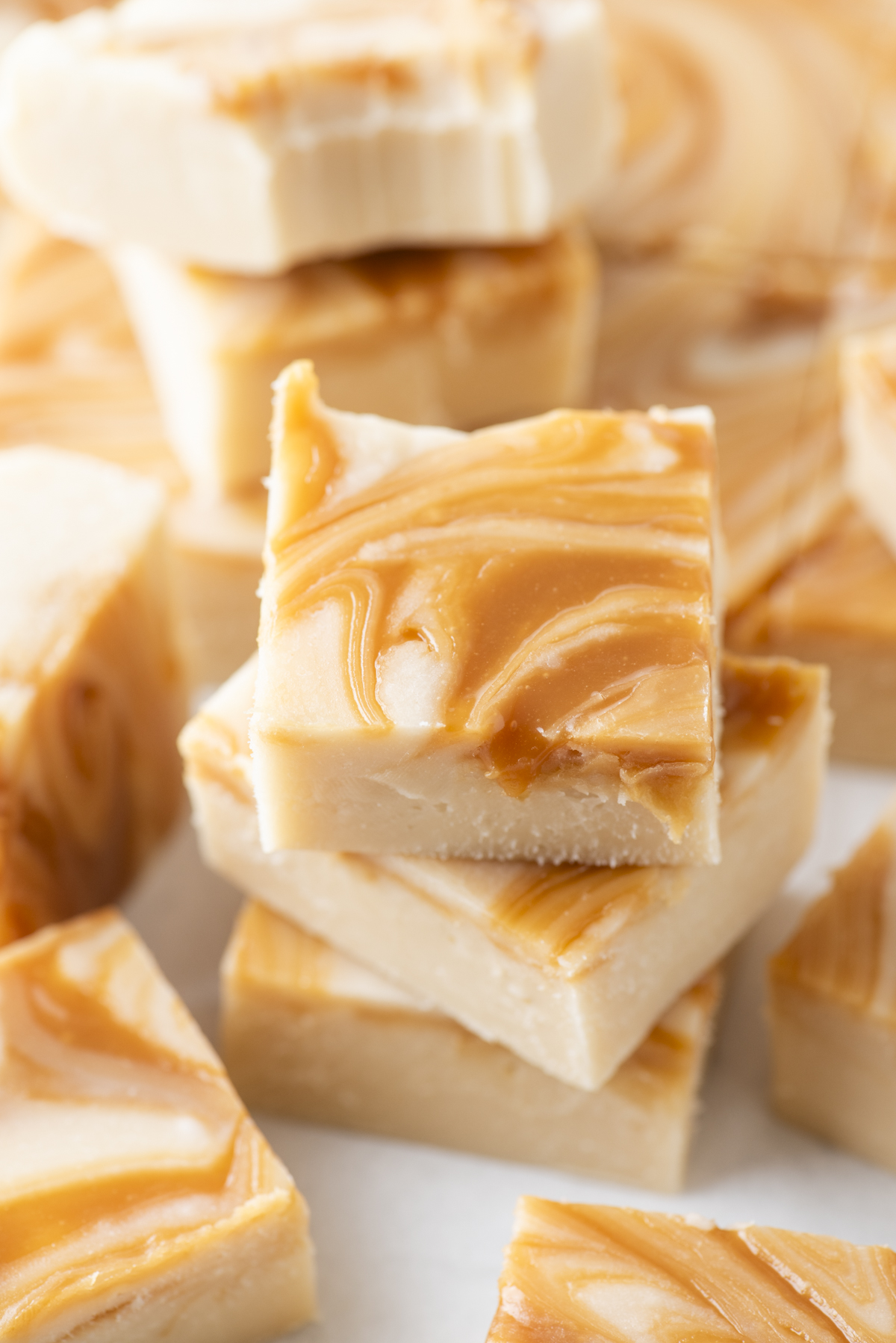 Squares of creamy white fudge topped with golden caramel swirls are stacked on each other, reminiscent of decadent caramel apple fudge, with more pieces blurred in the background. The surface appears smooth and glossy.