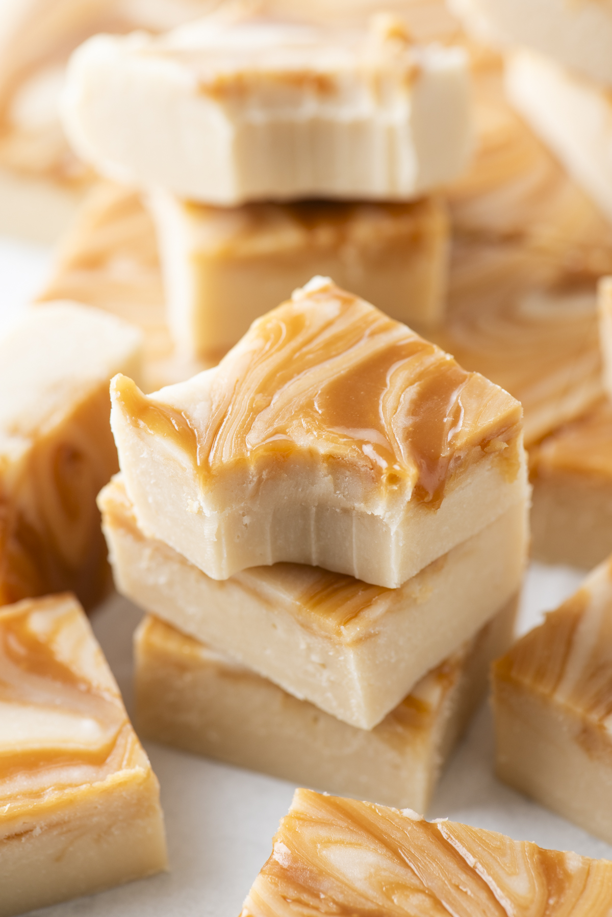 Squares of creamy caramel apple fudge with swirled caramel patterns are stacked, with one piece showing a bite taken out. The background is filled with more pieces of similar fudge.