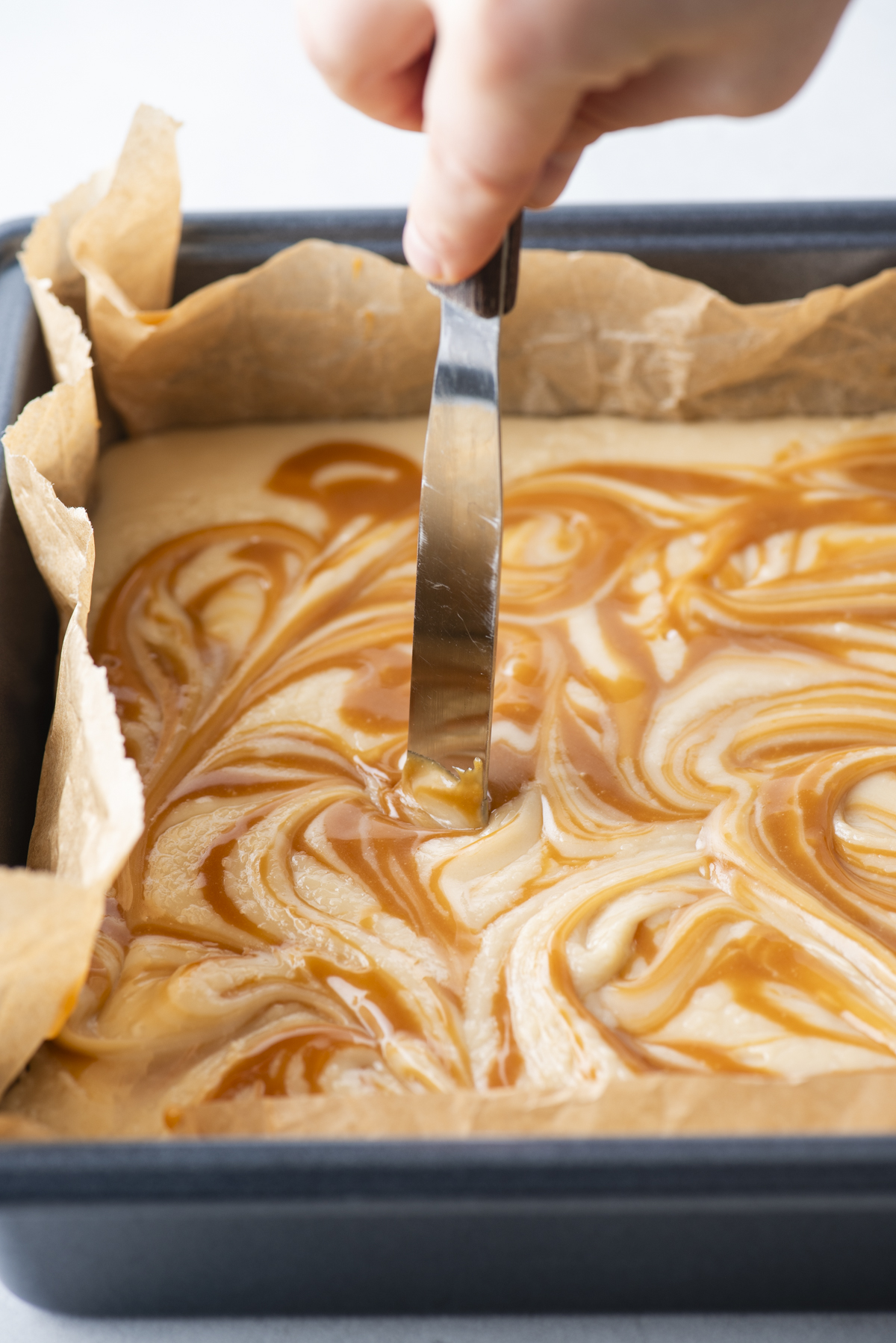 A hand swirls caramel apple fudge sauce into batter with a knife in a parchment-lined baking pan, creating a marbled effect.