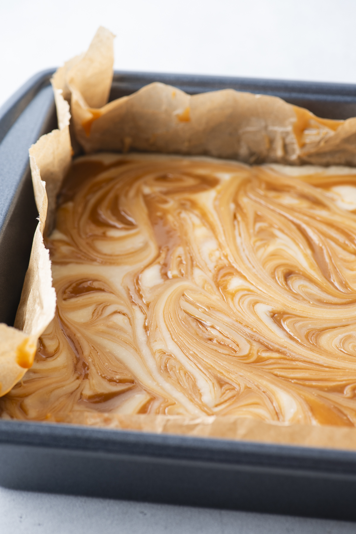 A close-up of a square baking pan lined with parchment paper, filled with a creamy caramel apple fudge batter swirled with caramel, ready to be baked.