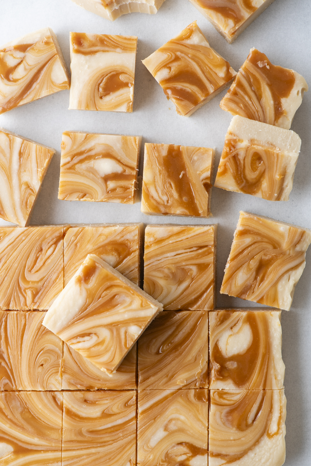 Squares of caramel apple fudge with caramel swirls are arranged on a white surface, some neatly cut and others scattered, showcasing their creamy texture and distinct marbled pattern.