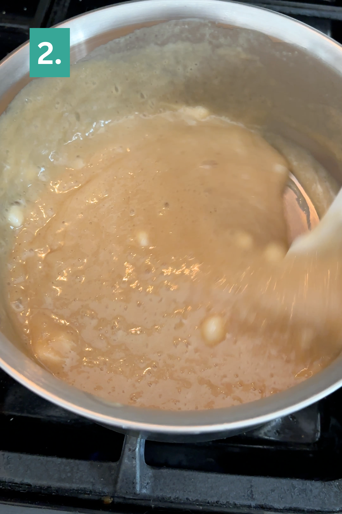 A pot on a stove with a thick beige mixture being stirred; small lumps are visible, suggesting the early stages of caramel apple fudge. A green box with the number 2 is in the top left corner.