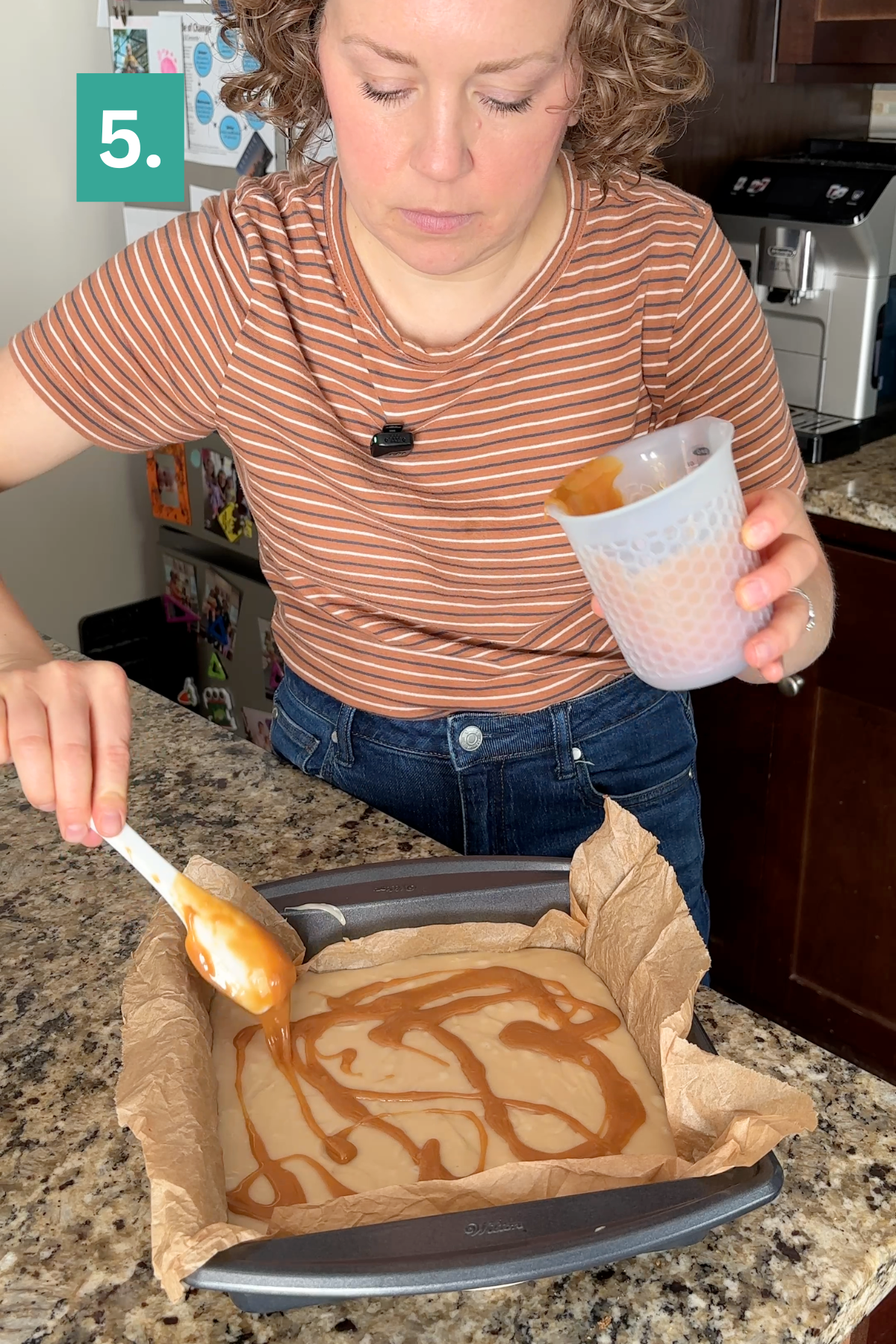 A woman pours caramel sauce over batter in a parchment-lined baking pan, preparing a decadent caramel apple fudge. She uses a spatula while holding a measuring cup, with a green label showing the number 5 in the top left corner.