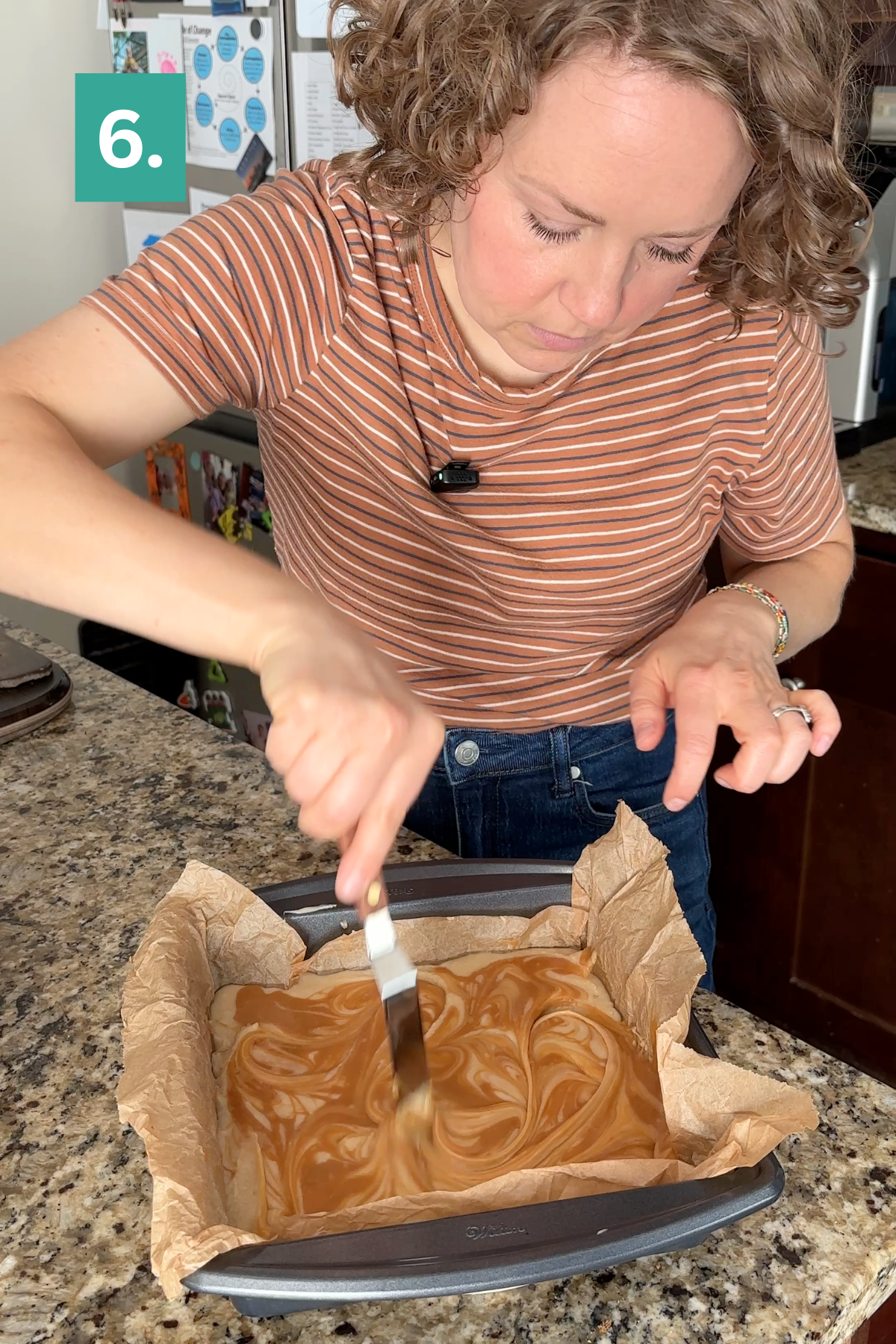 A person spreads a marbled caramel apple fudge batter in a square pan lined with parchment paper. A green box with the number 6 is in the upper left corner. The scene takes place in a kitchen.