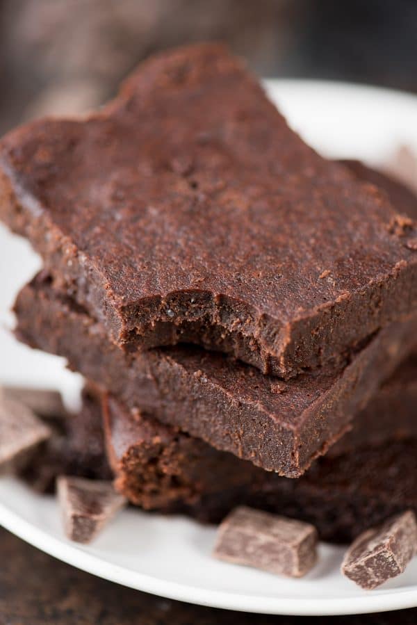 The healthier fudgy brownies of your dreams! No refined sugar, butter or oil, plus this recipe doesn’t call for any out of the ordinary ingredients. These are seriously fudgy and estimated to be 174 calories a slice! Three healthy fudgy brownies surrounded by small chocolate pieces on a white serving plate.