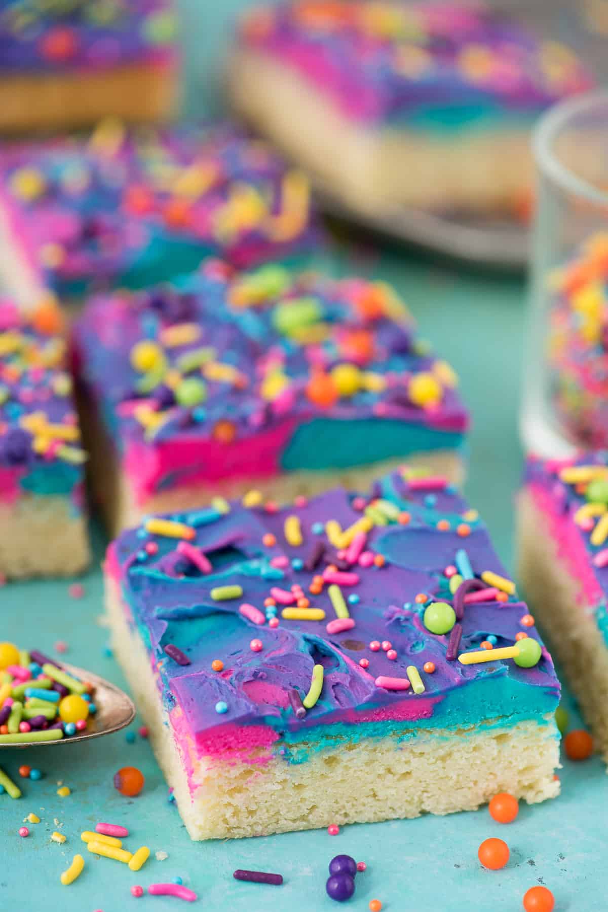 sugar cookie bars on a blue background with blue, pink and purple frosting and multi color sprinkles