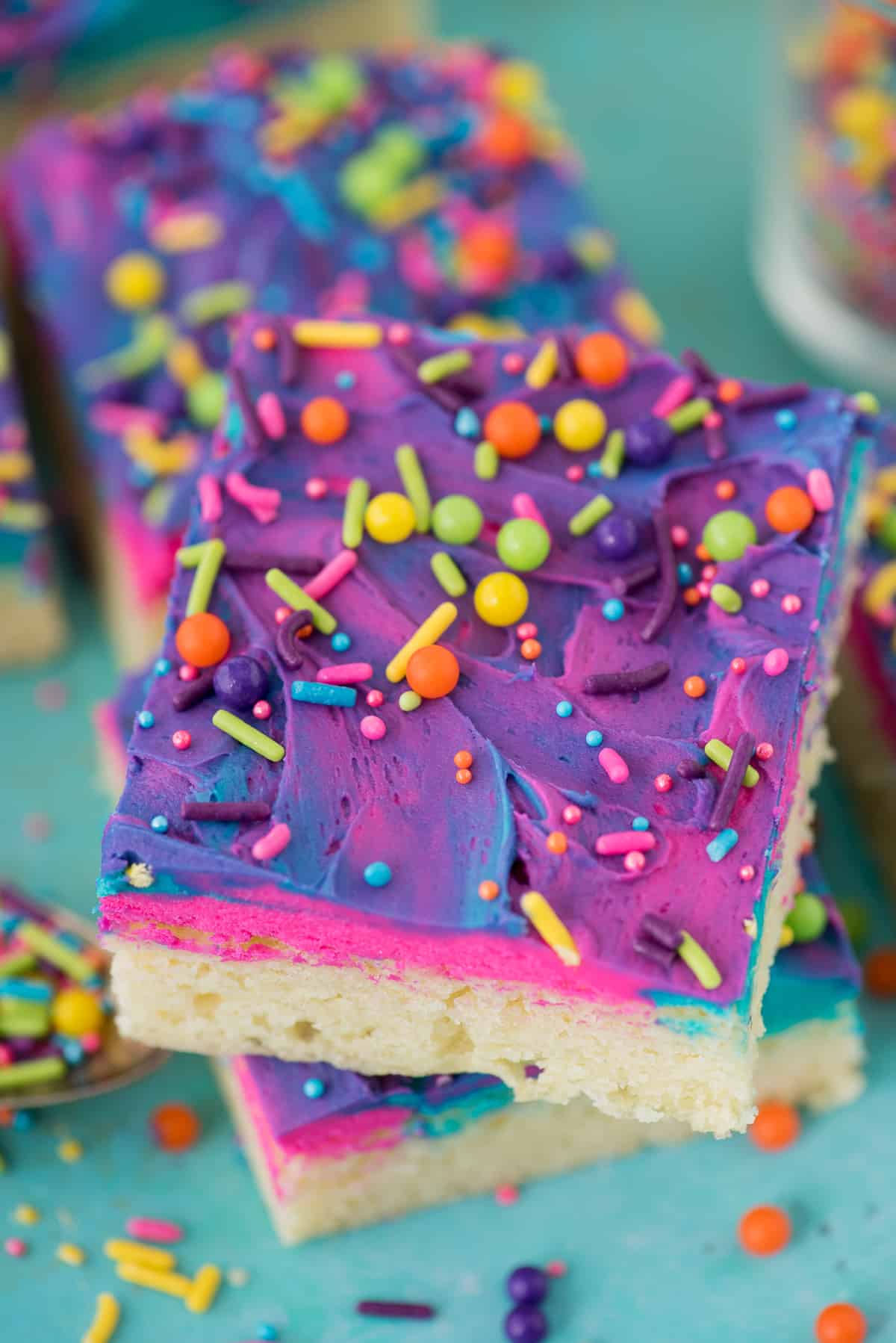 sugar cookie bars on a blue background with blue, pink and purple frosting and multi color sprinkles