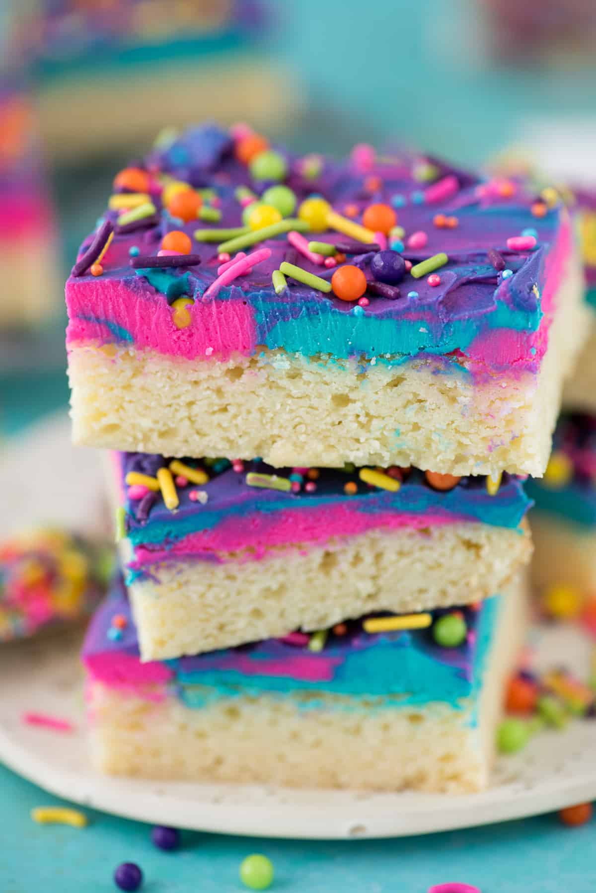 3 sugar cookie bars stacked on each other on a white plate with blue, pink and purple frosting and multi color sprinkles