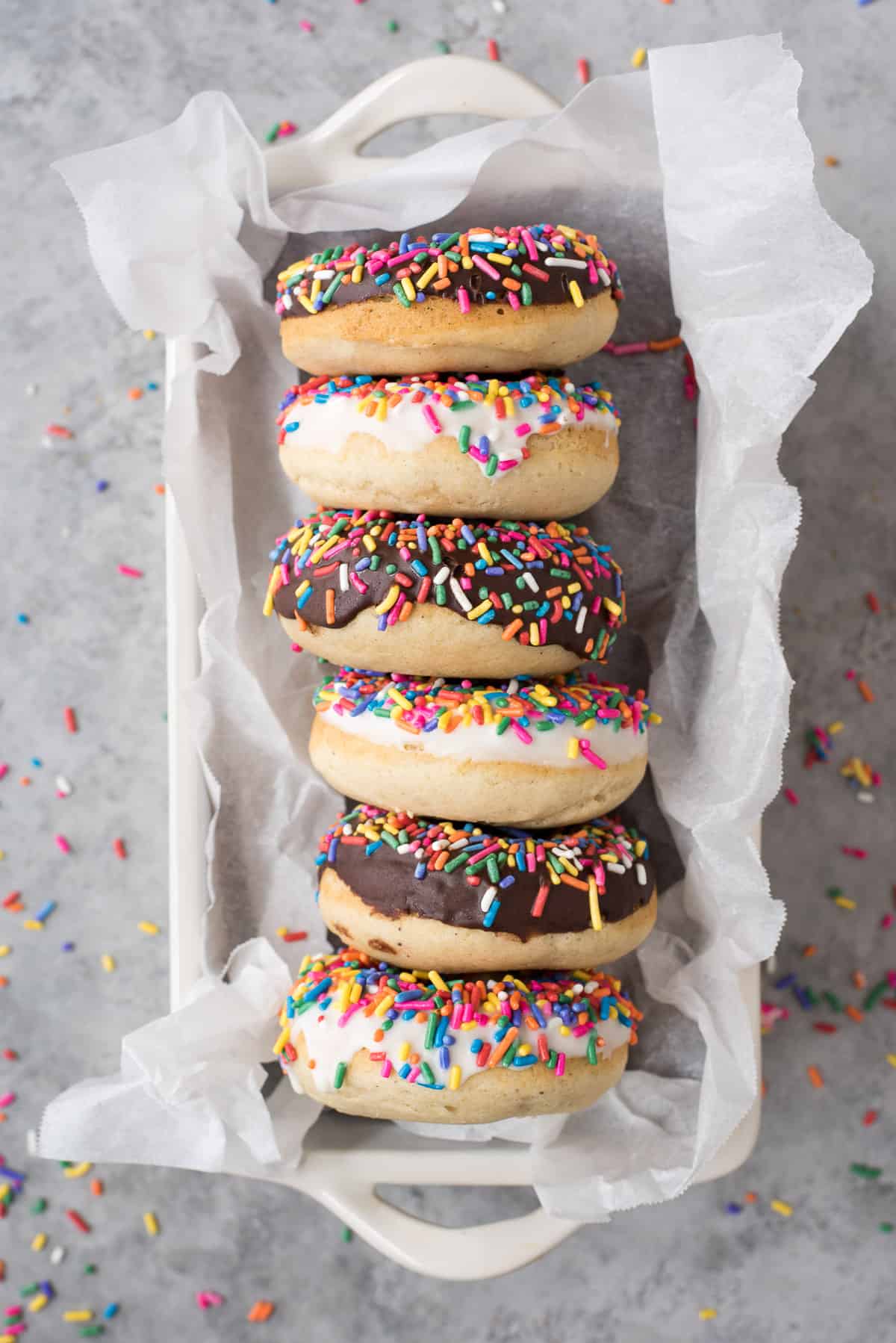 6 donuts with chocolate and white chocolate glaze with rainbow sprinkles lined up in white pan