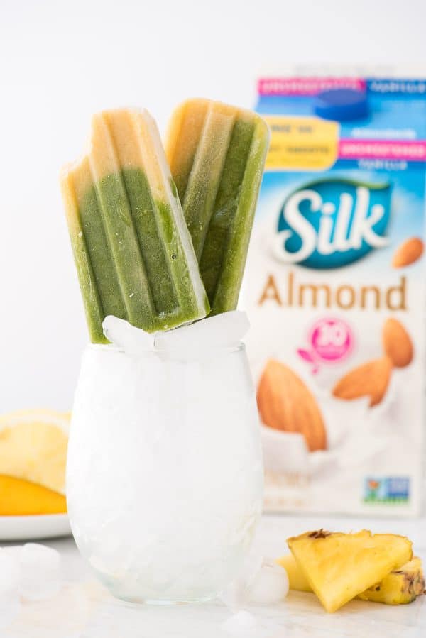 These Green Machine Popsicles are dairy free and customizable with different fruit options! Healthy popsicle recipe. #popsicles green spinach and mango popsicles propped up in a glass with ice. Container of silk almondmilk in the background.