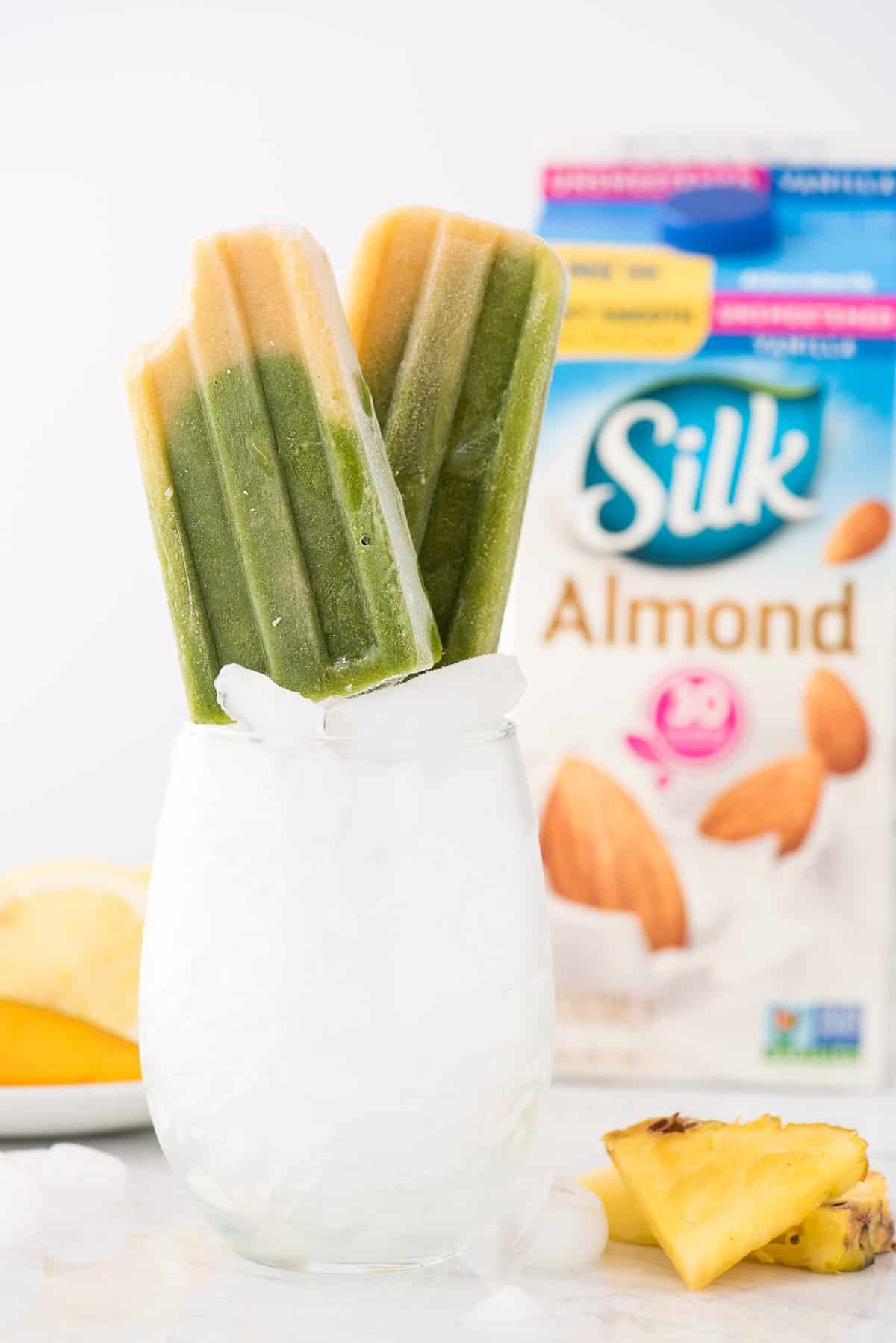 green spinach and mango popsicles propped up in a glass with ice. Container of silk almondmilk in the background. 