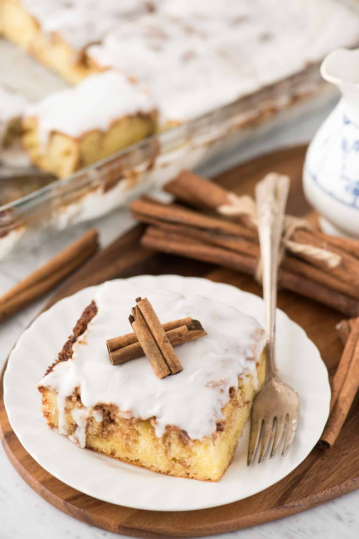 a piece of cinnamon swirl cake on white plate with white glaze on top and 2 cinnamon sticks