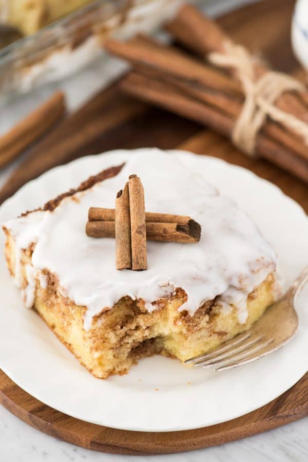 The most delicious doctored up HONEY BUN CAKE mix recipe! Easy to make with a box of cake mix, and with swirls of honey, cinnamon and brown sugar! Awesome breakfast cinnamon cake. a piece of cinnamon swirl cake on white plate with white glaze on top and 2 cinnamon sticks