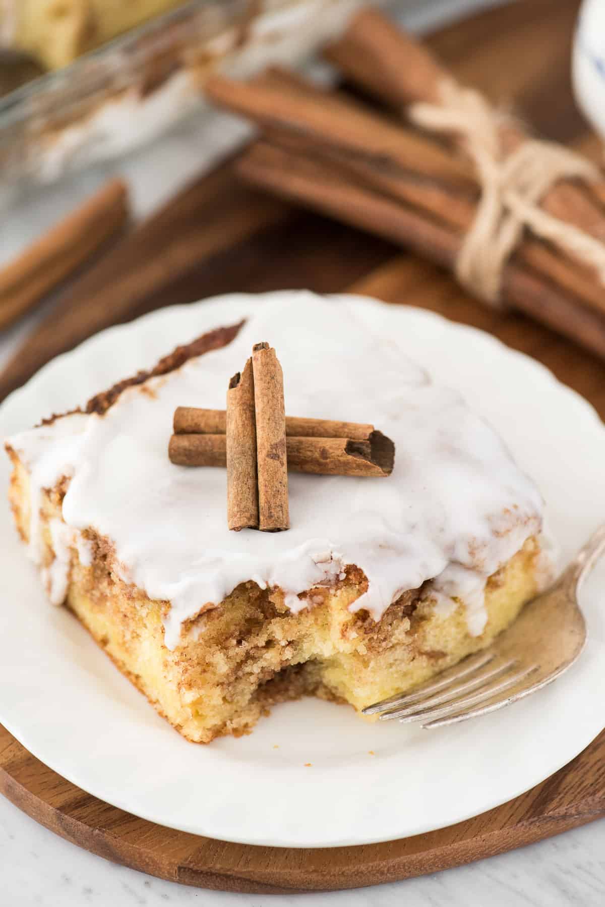 a piece of cinnamon swirl cake on white plate with white glaze on top and 2 cinnamon sticks