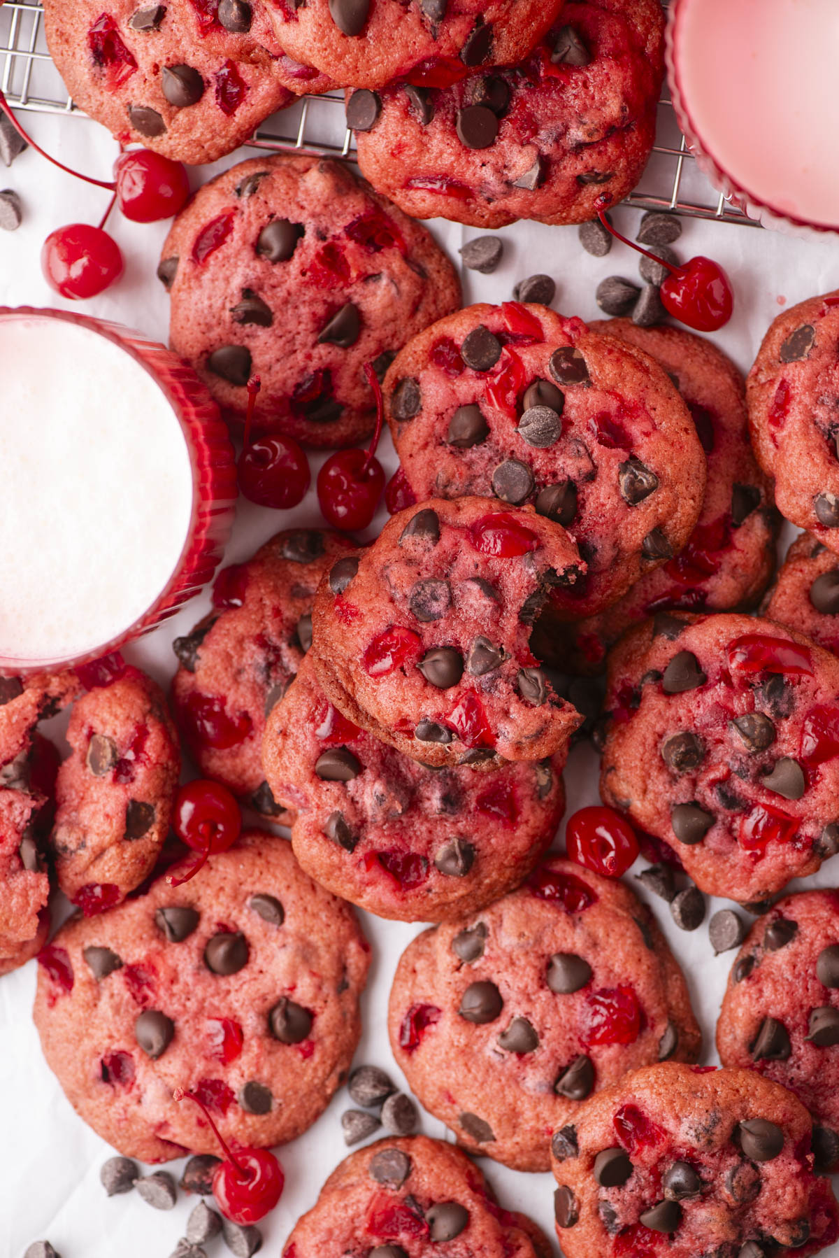 A close-up of pink cherry chocolate chip cookies scattered on a white surface, with chocolate chips and maraschino cherries visible. Two pink mugs, one filled with frothy milk, are placed among these delectable maraschino cherry cookies.