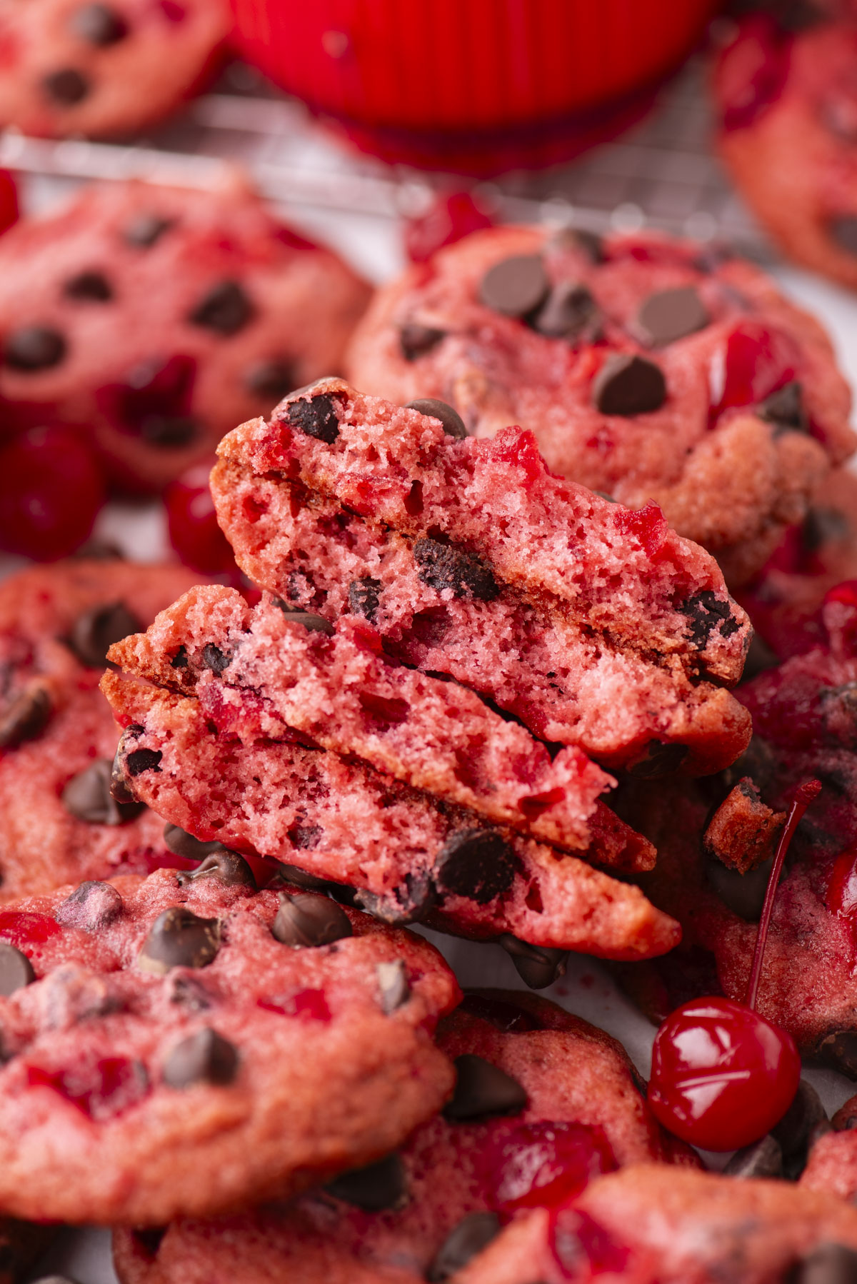 A close-up of cherry chocolate chip cookies, some broken in half to reveal their soft interior with melted chips and cherry pieces, surrounded by whole cherries and more cookies in the background.