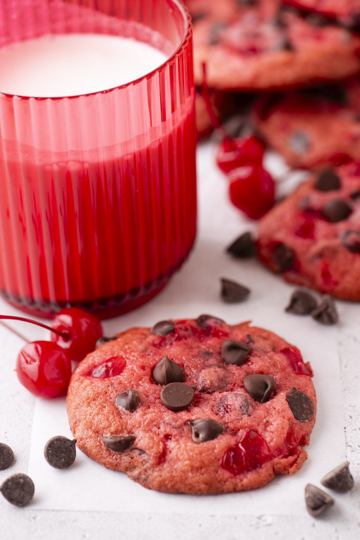 A close-up of a red cherry chocolate chip cookie on parchment paper, surrounded by chocolate chips and maraschino cherries, showcases the irresistible charm of maraschino cherry cookies. A red glass of milk sits in the background with other cookies slightly blurred behind.