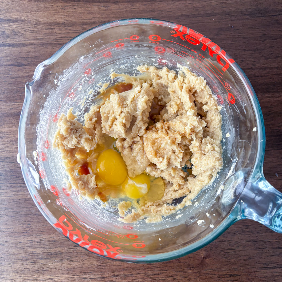 A glass Pyrex measuring cup on a wooden surface holds cookie dough mixture for cherry chocolate chip cookies, with two cracked eggs on top, ready to be mixed together.