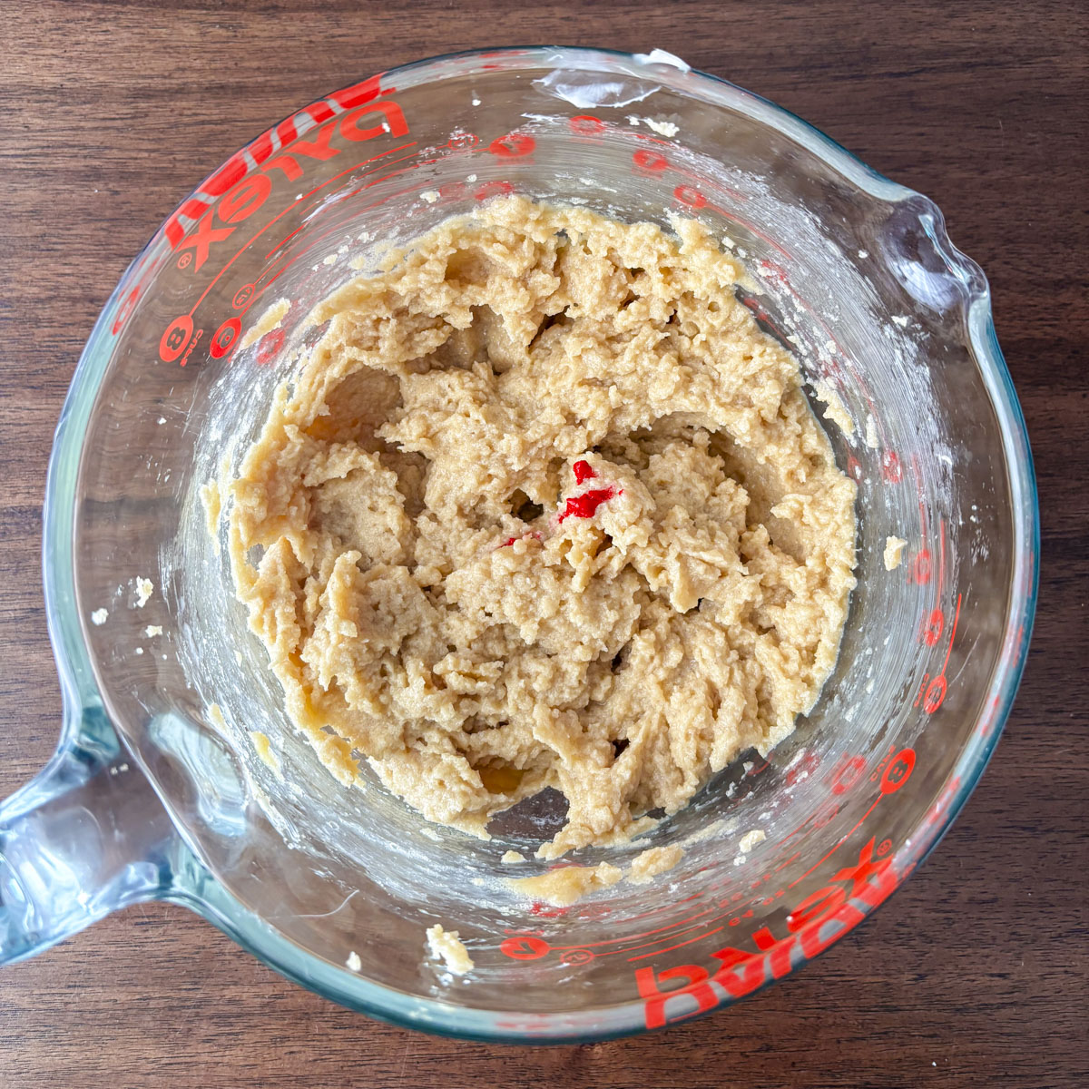 A glass Pyrex measuring cup filled with a mixed cherry cookie dough batter sits on a wooden table. The dough appears light brown and slightly chunky, with a few red bits—perfect for homemade maraschino cherry cookies.