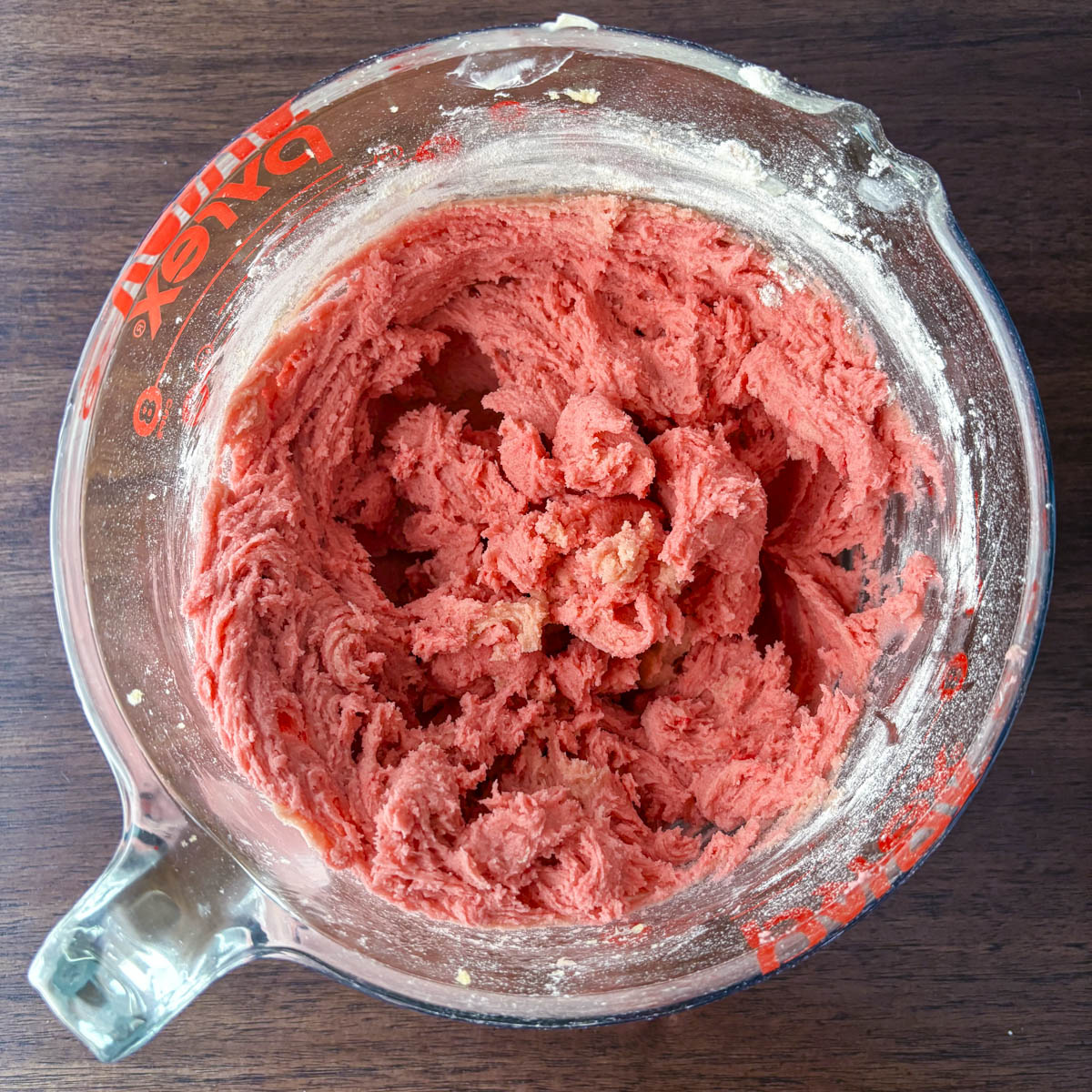 A glass mixing bowl on a dark wooden surface filled with bright pink maraschino cherry cookie dough, with some flour visible on the sides of the bowl.