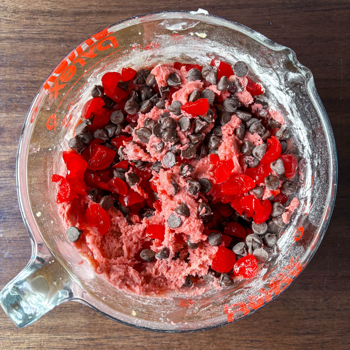 A glass measuring cup filled with pink cherry chocolate chip cookie dough, chocolate chips, and chopped red candied cherries sits on a wooden surface.