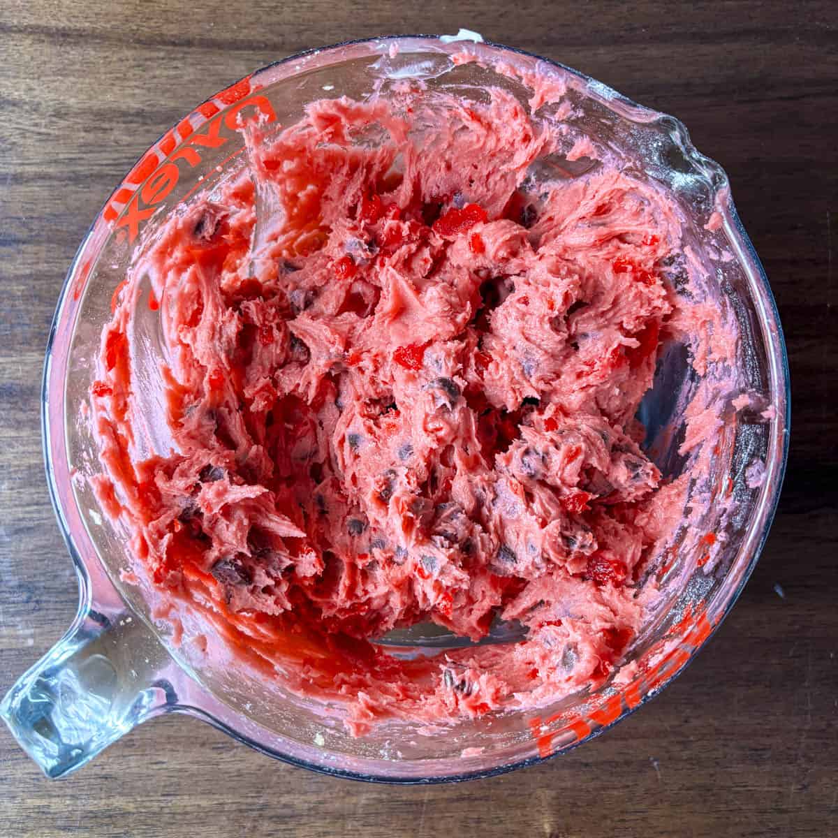 A glass mixing bowl filled with bright pink cherry chocolate chip cookie dough sits on a wooden surface, with bits of dough clinging to the sides.