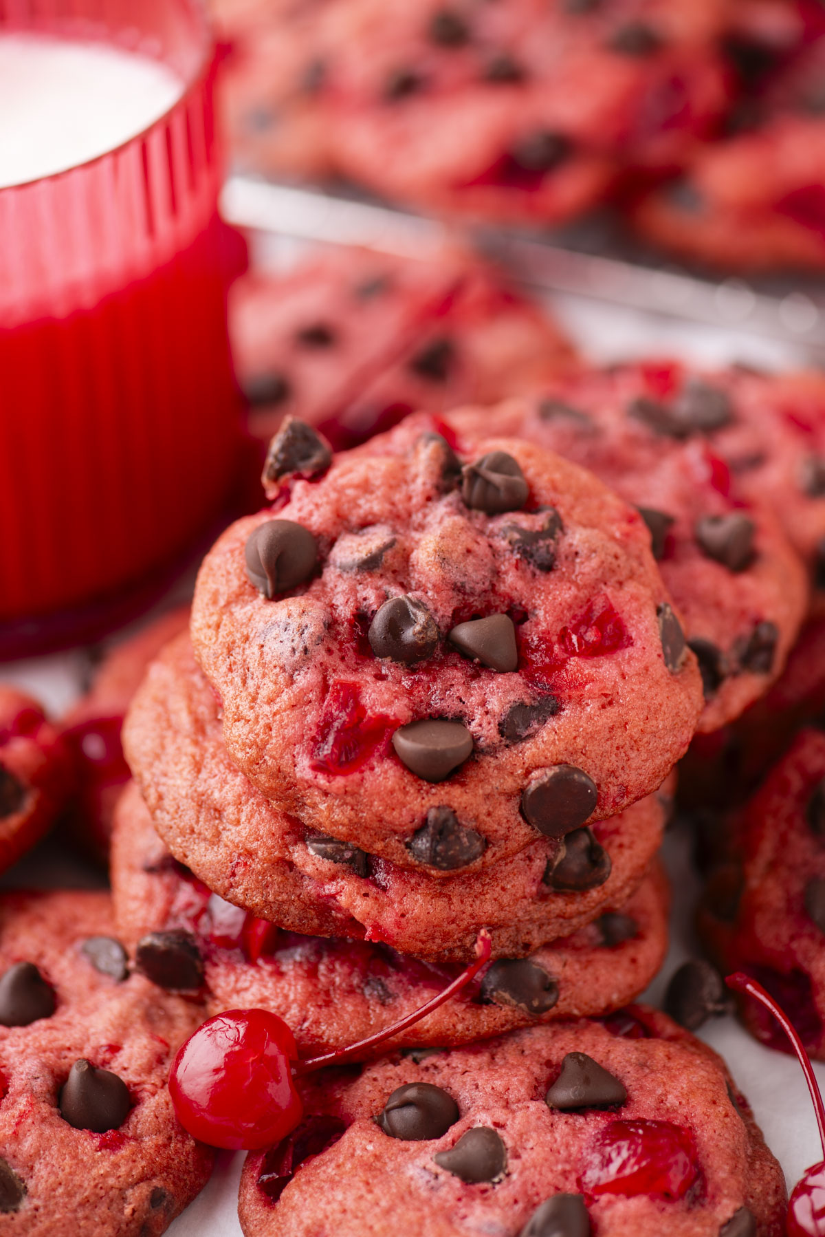 A stack of pink maraschino cherry cookies with chocolate chips and cherry pieces, topped with a maraschino cherry, sits beside a red cup of milk. More cherry chocolate chip cookies are visible in the background.
