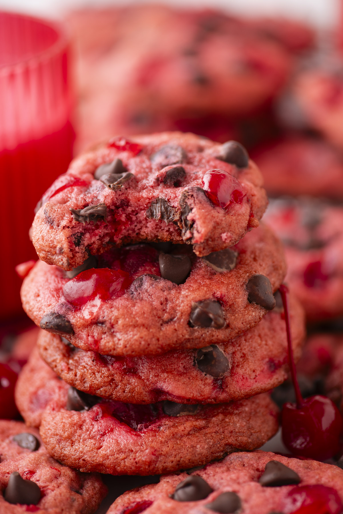 A stack of pink cherry chocolate chip cookies with visible cherries, topped with a bitten cookie, sits in front of a red container. Cherries and more cookies are scattered around.