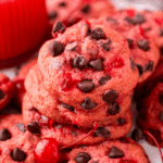 A close-up of stacked cherry chocolate chip cookies with pieces of cherries, surrounded by more cookies and a whole cherry in the foreground.