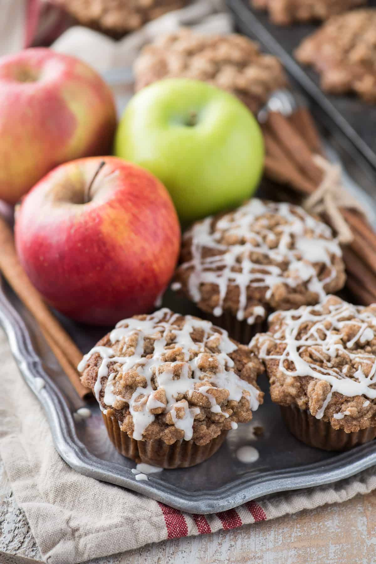applesauce spice muffins with crumb topping and white glaze on metal plate and apples in the background