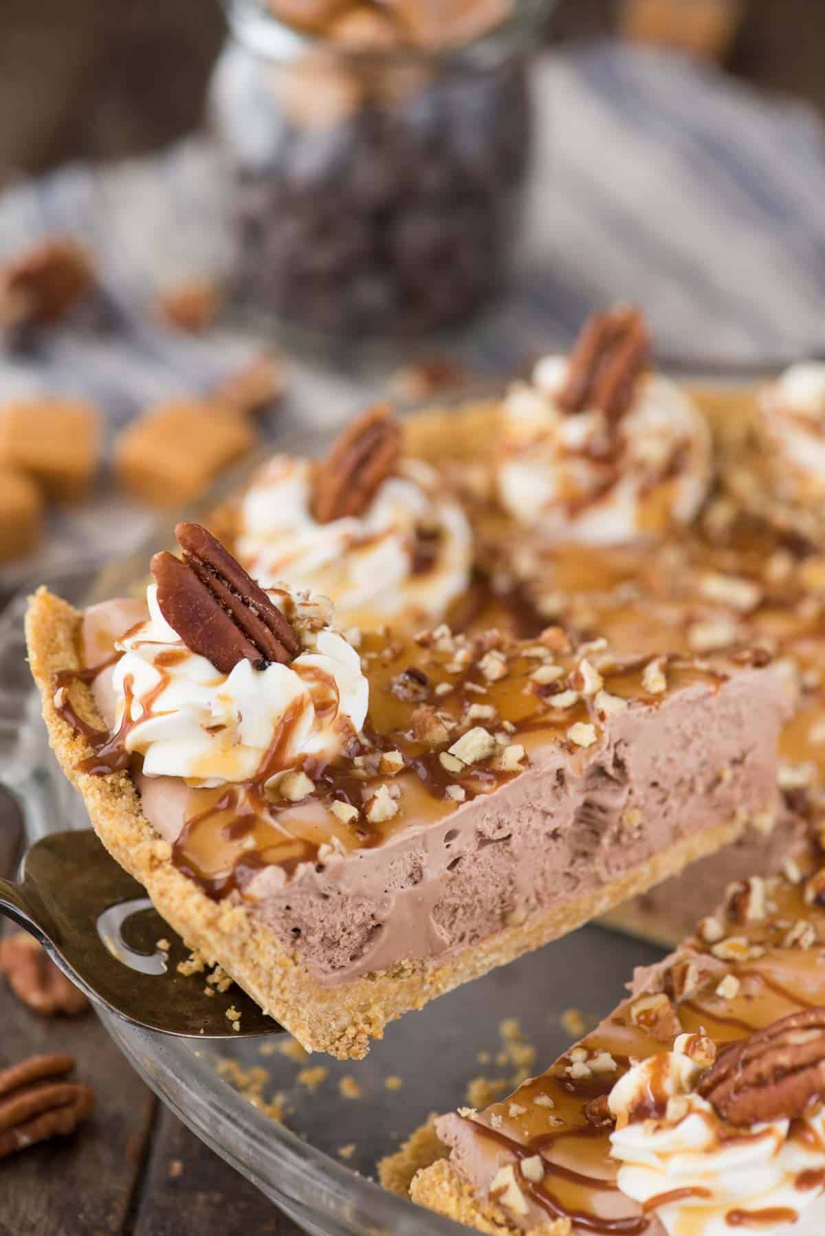 slice of turtle pie with chocolate filling, chopped pecans and caramel drizzle being removed from pie pan