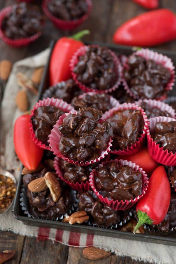 Crock pot or stove top spicy nut clusters! These dark chocolate cayenne nut clusters are popular during the holiday season. Plus there’s notes for making them less spicy or packed with heat! dark chocolate cayenne nut clusters in red mini muffin paper cups on wood background