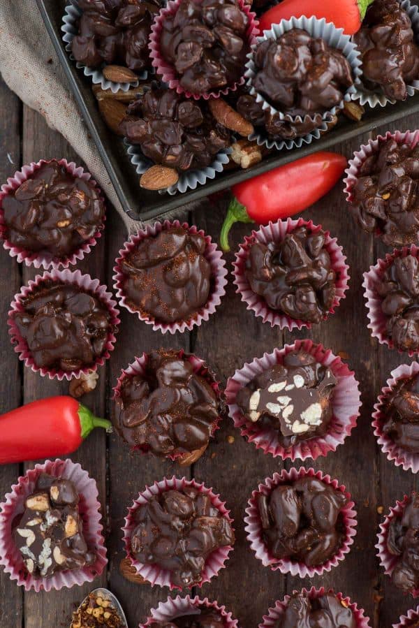 Crock pot or stove top spicy nut clusters! These dark chocolate cayenne nut clusters are popular during the holiday season. Plus there’s notes for making them less spicy or packed with heat! dark chocolate cayenne nut clusters in red mini muffin paper cups on wood background
