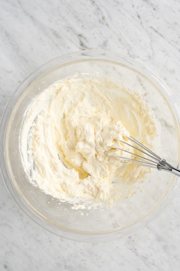 How to make stabilized cheesecake flavored whipped cream that you can use on cake, cupcakes, pies, and as fruit dip! Imagine this cheesecake flavored whipped cream on strawberry pie! #whippedcream #flavoredwhippedcream #cheesecakewhippedcream cheesecake whipped cream in glass bowl with whisk