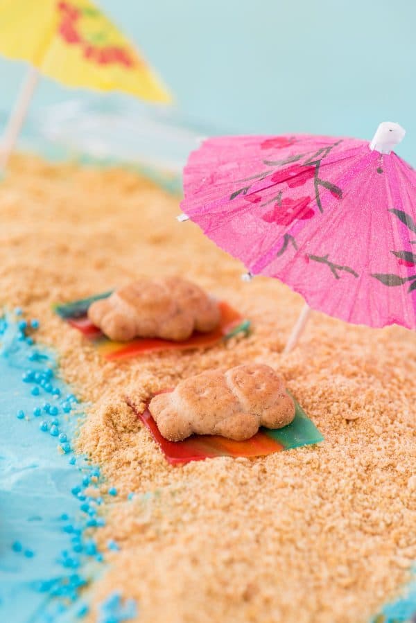 This beach theme poke cake would be a hit at a beach theme party or under the sea theme party! Kids will have fun decorating the top of this beach cake with teddy grahams, gummy sea creatures, and graham cracker sand. #beachpokecake #pokecake #beachcake beach theme cake, teddy grahams on fruit roll up towels sitting on graham cracker sand under a toothpick umbrella