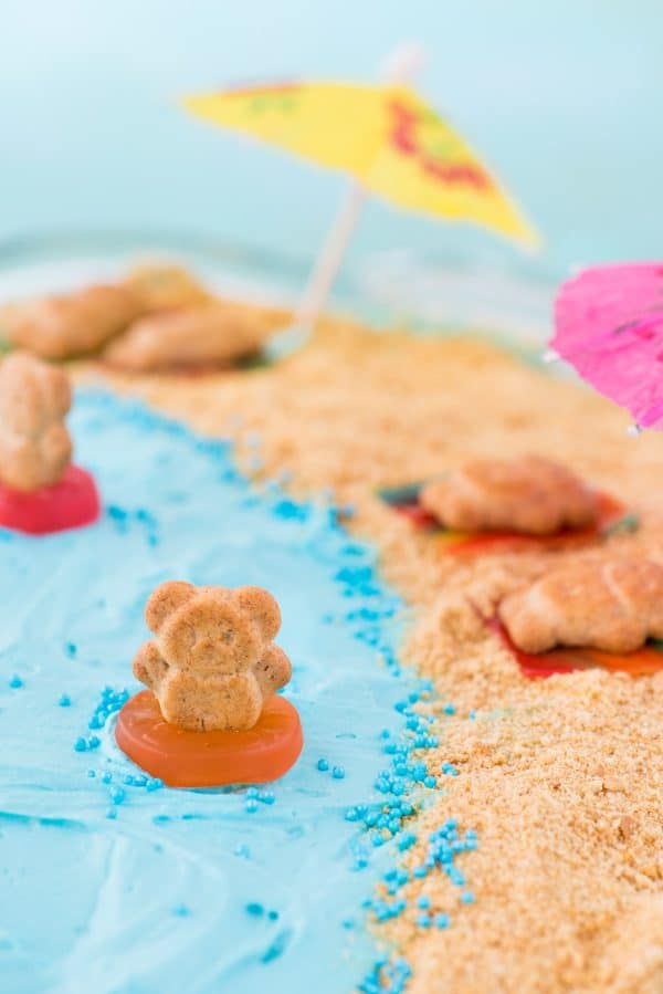 This beach theme poke cake would be a hit at a beach theme party or under the sea theme party! Kids will have fun decorating the top of this beach cake with teddy grahams, gummy sea creatures, and graham cracker sand. #beachpokecake #pokecake #beachcake beach theme cake, teddy graham in gummy life saver swimming