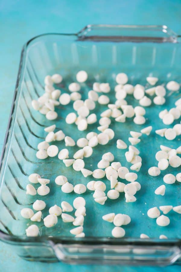 This funfetti earthquake cake is a white cake mix loaded with sprinkles and funfetti chocolate chips. Swirl in the cream cheese frosting layer and it bakes into the cake creating crevices and cracks on top of the cake. #earthquakecake #funfetticake #cake white chocolate chips in bottom of glass 9x13 inch pan