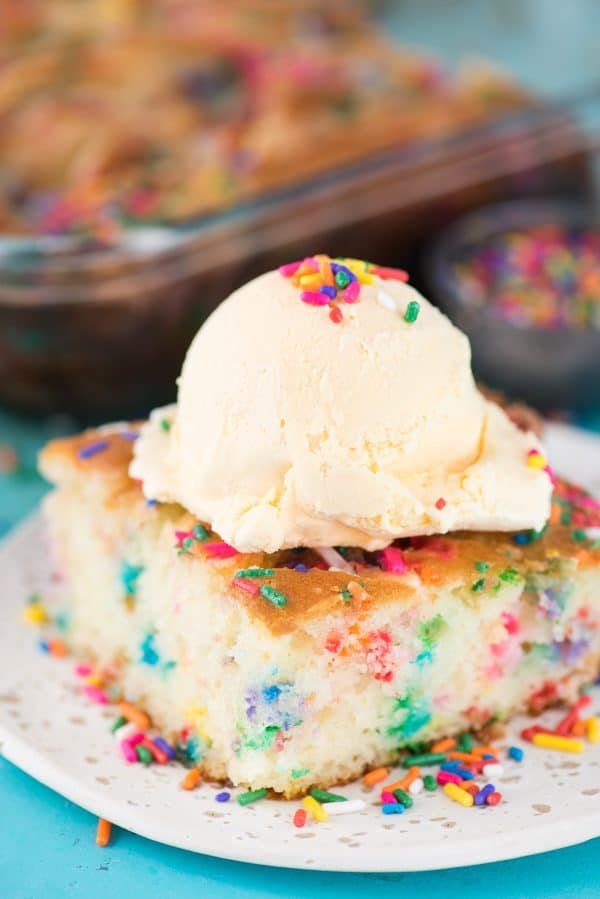 This funfetti earthquake cake is a white cake mix loaded with sprinkles and funfetti chocolate chips. Swirl in the cream cheese frosting layer and it bakes into the cake creating crevices and cracks on top of the cake. #earthquakecake #funfetticake #cake slice of funfetti earthquake cake with scoop of vanilla ice cream on white plate on blue background