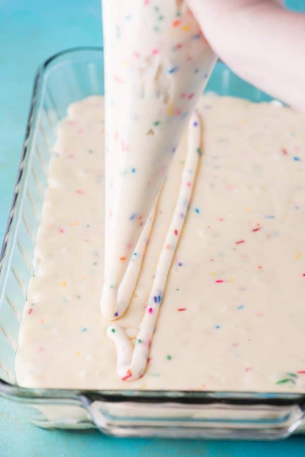 This funfetti earthquake cake is a white cake mix loaded with sprinkles and funfetti chocolate chips. Swirl in the cream cheese frosting layer and it bakes into the cake creating crevices and cracks on top of the cake. #earthquakecake #funfetticake #cake piping cream cheese filing on top of white cake mix in glass 9x13 inch pan