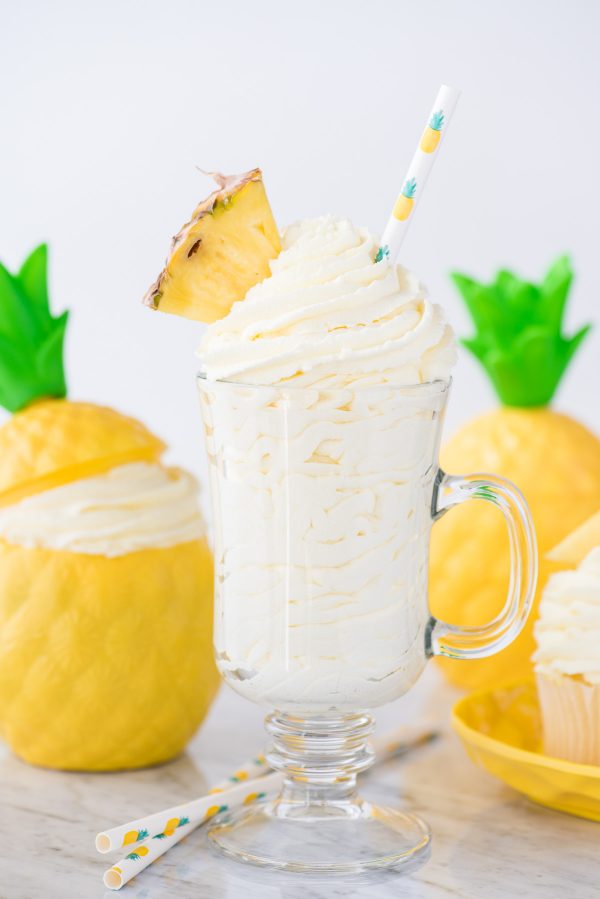Easy to make 3 ingredient pineapple whipped cream frosting! This pineapple whipped cream makes a great frosting for cake & cupcakes. Use it as a whipped topping on tropical drinks too! #whippedcream #pineapplefrosting #pineapplewhippedcream pineapple whipped cream in glass cup with a pineapple piece and pineapple printed straw