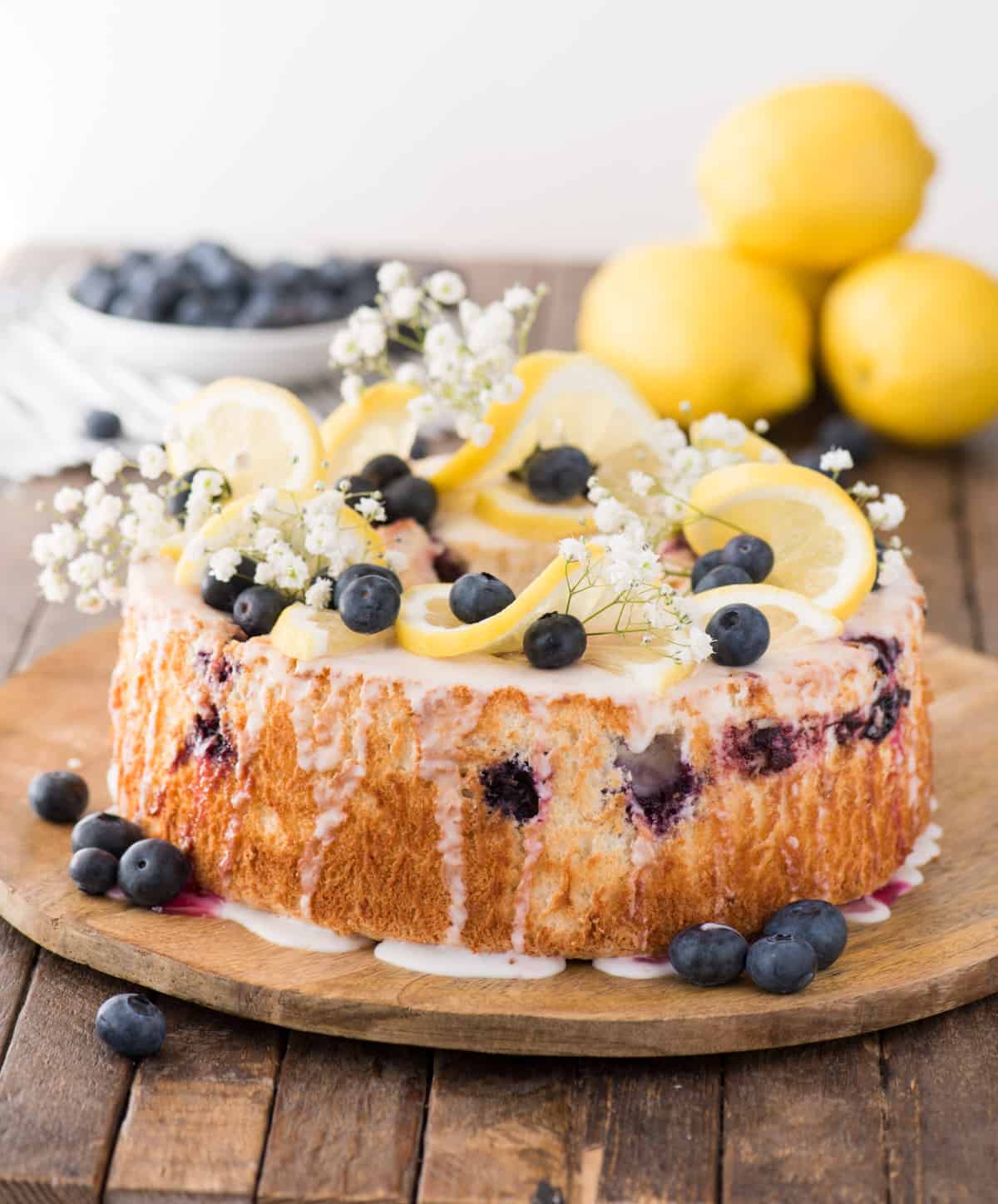 Lemon blueberry angel food cake with lemon slices, blueberries and baby’s breath as garnish on top of the cake on a wood serving plate. 