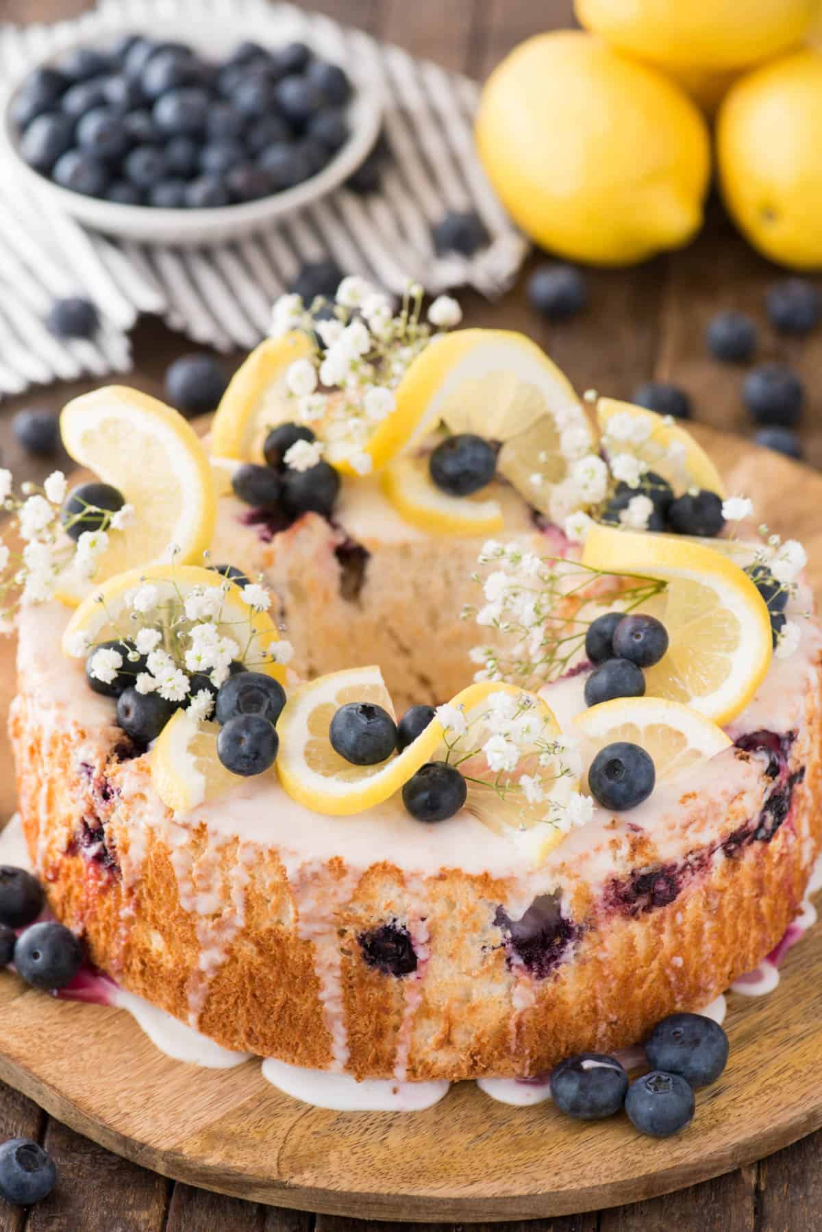 Lemon blueberry angel food cake with lemon slices, blueberries and baby’s breath as garnish on top of the cake on a wood serving plate. 