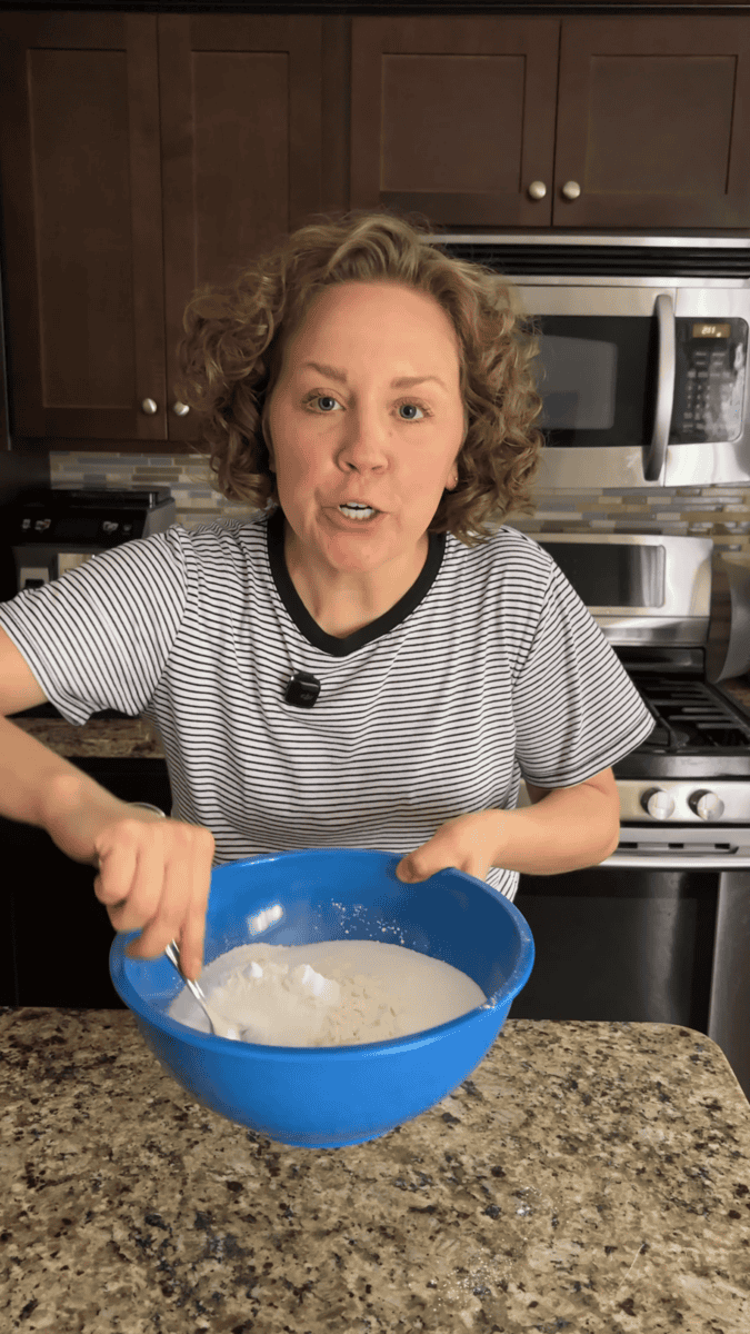 A person with curly hair mixes ingredients for coconut chocolate chip muffins in a blue bowl on a kitchen counter, wearing a striped shirt, with an oven and microwave in the background.
