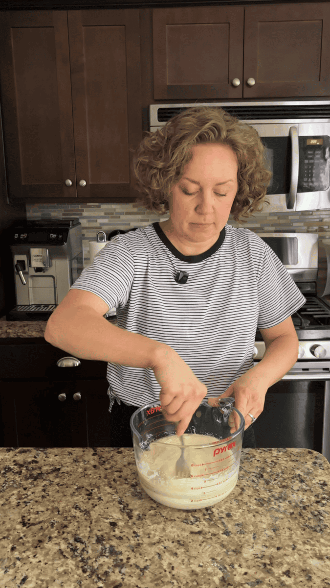 A person with curly hair in a striped shirt mixes batter for coconut chocolate chip muffins in a glass measuring cup in a kitchen with dark wood cabinets and stainless steel appliances.