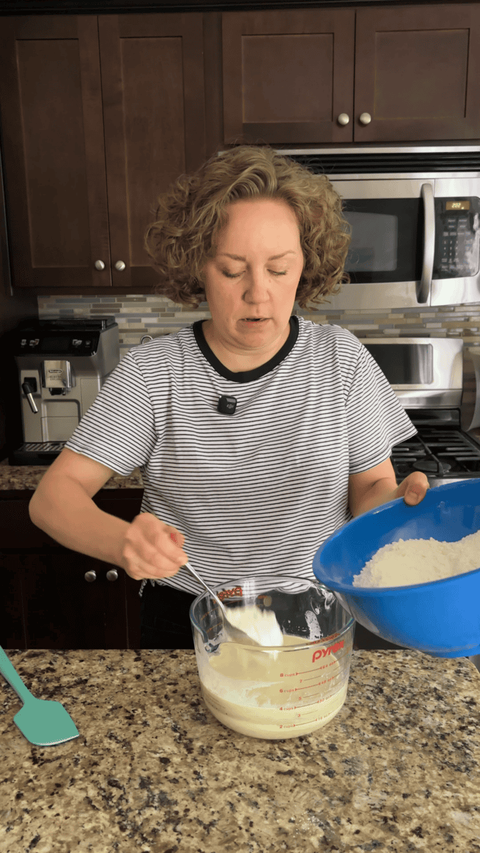 A person stands in a kitchen, holding a blue bowl of flour and spooning it into a glass mixing cup containing a white mixture, preparing ingredients for coconut chocolate chip muffins on a granite countertop.